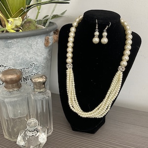 May include: A multi-strand pearl necklace with a crystal accent and matching pearl earrings. The necklace is displayed on a black stand.