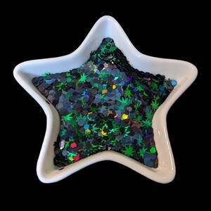 May include: A white star-shaped dish filled with iridescent green, black, and rainbow glitter. The glitter is shaped like leaves and hexagons.