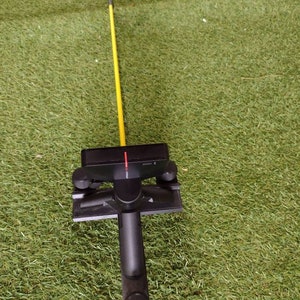 May include: A black golf swing training aid with a yellow alignment rod. The device has a handle, a rectangular base, and adjustable knobs. The alignment rod is thin and extends upwards.