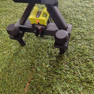 May include: A black tripod with adjustable knobs supports a yellow and black Ryobi laser level. A red laser line projects onto the green grass. The setup is designed for leveling and alignment tasks.
