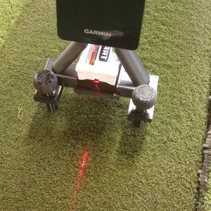 May include: A black Garmin device with a red laser beam pointing downwards. The device is mounted on a tripod with two black legs and a white box.