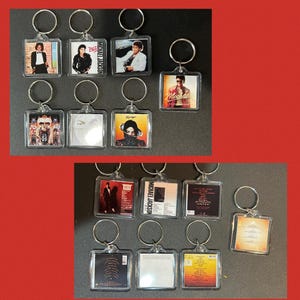 May include: A collection of square acrylic keychains featuring various Michael Jackson album covers and back tracklists. Each keychain has a silver metal split ring attached to the top. The designs include portraits of the artist and text.