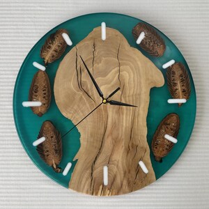 May include: Round wall clock with a teal resin background and a natural wood center. The clock features black hands, white hour markers, and decorative seed pods. A unique, handcrafted home decor item.
