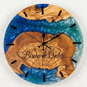 Puede incluir: Un reloj de pared redondo hecho de una rodaja de madera natural con un patrón de veta prominente, con una incrustación de resina epoxi azul y turquesa. La esfera tiene marcadores de hora negros, manecillas negras y el texto Bircan & Önder.