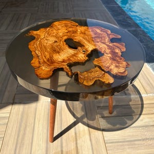 Könnte beinhalten: Ein runder Couchtisch mit einer transparenten, glänzenden Oberfläche. Die Tischplatte zeigt ein einzigartiges, organisches Holzmaserungsmuster, das in dunklem Harz eingeschlossen ist. Er steht auf drei Holzbeinen und schafft eine moderne, natürliche Ästhetik.