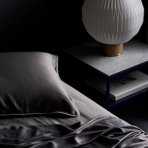 May include: A dark gray silk sheet with a white piping detail, a matching pillow, and a white pleated lampshade on a bedside table.