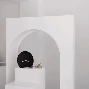 Peut inclure: Une horloge ronde noire avec des aiguilles dorées est posée sur un piédestal blanc devant une arche blanche.