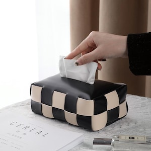 May include: A black and cream woven tissue box with a hand taking a white tissue. The box is rectangular and is on a table next to a magazine with the word "CEREAL" on the cover.