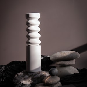 May include: A white ceramic vase with a unique, segmented design sits on a gray rock. The vase is surrounded by other gray rocks and black fabric.