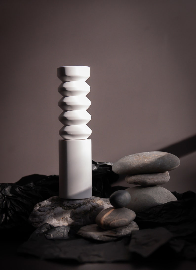 May include: A white ceramic vase with a unique, segmented design sits on a gray rock. The vase is surrounded by other gray rocks and black fabric.
