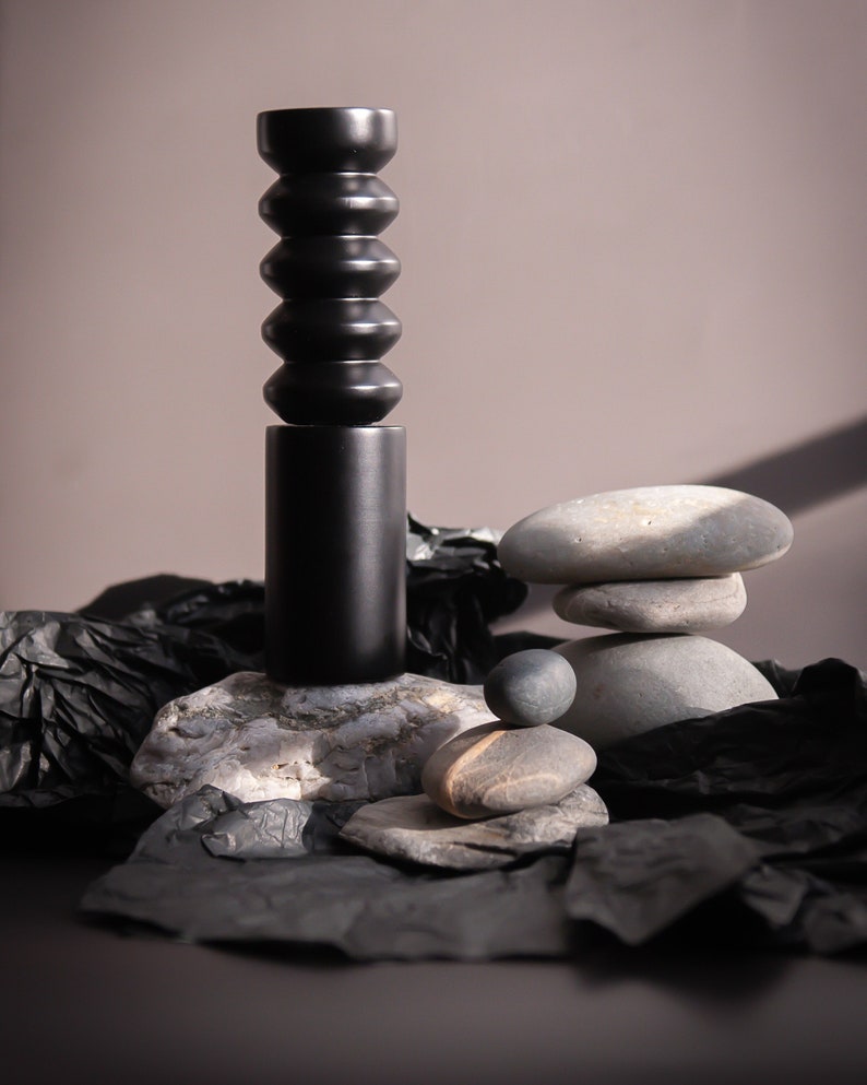 May include: A black ceramic vase with a textured, segmented design sits on a gray rock. The vase is surrounded by gray rocks and black crumpled paper.