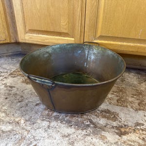 May include: A weathered, antique copper bucket with two small handles and a rounded shape. The bucket has a dark patina and sits on a mottled, light-colored surface. The background features wooden cabinetry.