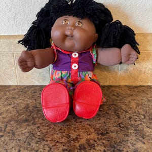May include: A vintage Cabbage Patch Kid doll with dark skin, black yarn hair, and brown eyes. The doll wears a purple top with white buttons and a colorful floral print dress. Red shoes complete the look.