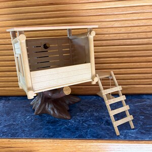 May include: A miniature wooden treehouse with a ladder, perched on a brown tree trunk base. The treehouse has a light-colored roof and walls with horizontal slats. The ladder is made of small wooden rungs and string.
