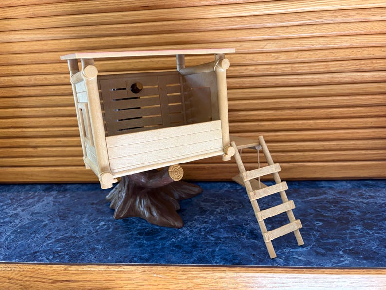 May include: A miniature wooden treehouse with a ladder, perched on a brown tree trunk base. The treehouse has a light-colored roof and walls with horizontal slats. The ladder is made of small wooden rungs and string.