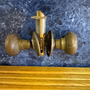 May include: An antique brass door knob set with a mortise lock. The set includes two rounded knobs, a central lock mechanism, and two circular plates. The brass has a weathered appearance, suggesting age and use. The background is a dark, marbled surface.