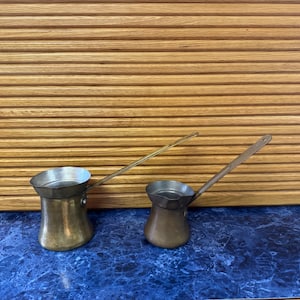 May include: Two antique brass coffee pots with long handles. The pots have a classic, rounded shape and are made of aged brass. They sit on a blue countertop with a wooden slatted background. The pots are of different sizes.