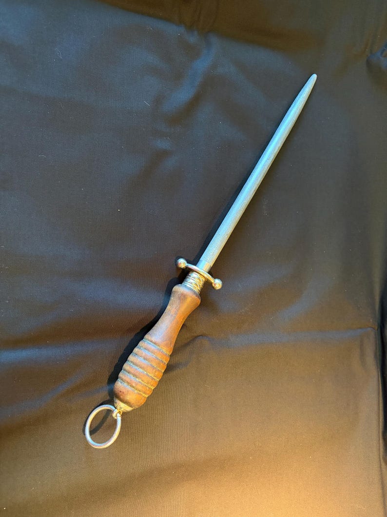 May include: A steel sharpening rod with a tapered, textured gray metal shaft and a cylindrical wooden handle. The handle features horizontal grooves, a metal bolster with a small guard, and a circular metal ring attached to the base for hanging.
