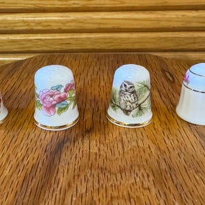 May include: Four white porcelain thimbles with gold trim are displayed on a wooden surface. Each thimble features a different design: a bird, pink flowers, an owl, and pink roses. The thimbles are decorative and collectible.