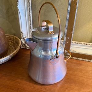 May include: A vintage metal teapot with a copper finish. The teapot has a rounded body, a spout, and a hinged lid. It features a copper handle and a small copper handle on the lid. The teapot is sitting on a wooden surface.