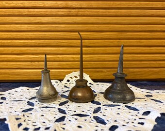 May include: Three vintage metal oil cans with long, pointed spouts. The left can is silver-toned, the center can is a warm copper color, and the right can is a dark, weathered gray. Each features a rounded base and a narrow, upright nozzle.