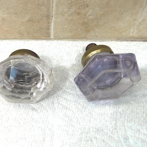 May include: Two octagonal glass cabinet knobs with faceted surfaces and brass-toned metal bases. One knob is clear glass, while the other displays a light purple tint. Both feature a multi-sided geometric shape with a central mounting point.