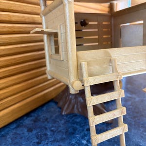 May include: A tan-colored toy treehouse with a ladder, window, and a small roof. The treehouse is made of plastic and has a wooden appearance. The treehouse sits on a brown base and is on a blue surface.