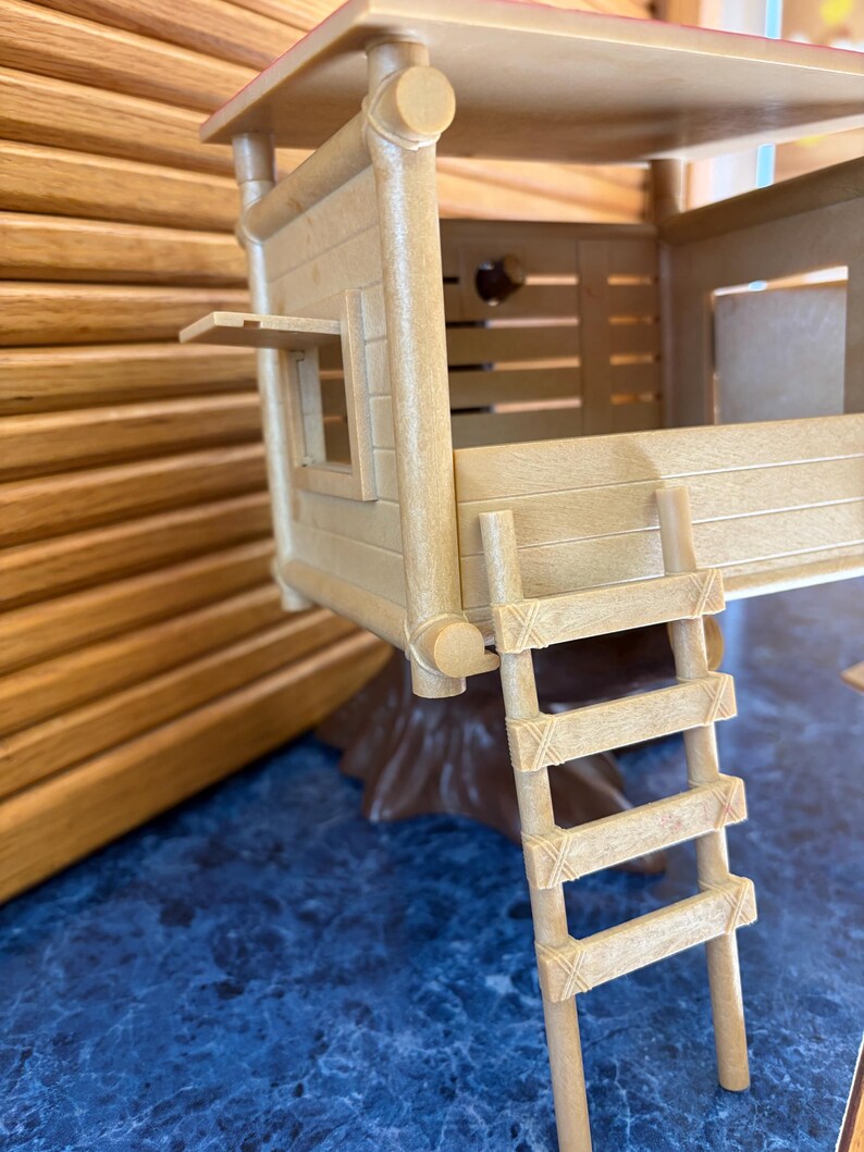 May include: A tan-colored toy treehouse with a ladder, window, and a small roof. The treehouse is made of plastic and has a wooden appearance. The treehouse sits on a brown base and is on a blue surface.