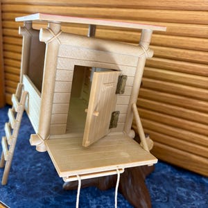 May include: A tan-colored miniature treehouse with a ladder, a swing, and an open door. The treehouse has a red-trimmed roof and is set on a brown base. The treehouse is made of wood-like material.