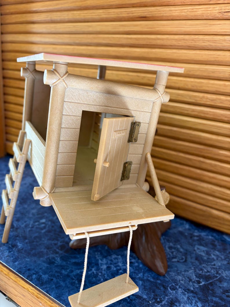 May include: A tan-colored miniature treehouse with a ladder, a swing, and an open door. The treehouse has a red-trimmed roof and is set on a brown base. The treehouse is made of wood-like material.