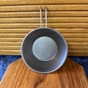 May include: A small, round, metal pan with a handle, resting on a wooden cutting board. The pan has a circular base and a raised rim. The handle is a simple, curved design. The background features a wooden surface.