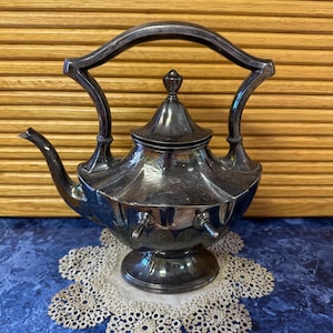 May include: A silver teapot with a decorative handle and a domed lid. The teapot is sitting on a white lace doily.