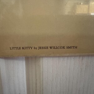 May include: A framed print with the text "LITTLE KITTY by JESSIE WILLCOX SMITH" in a serif font. The print has a light brown background and is set against a white, vertically-paneled wall.