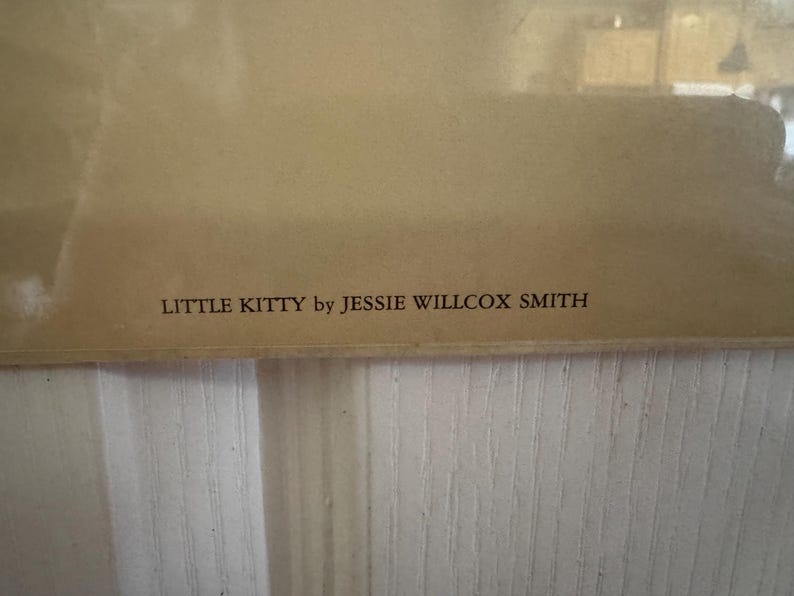 May include: A framed print with the text "LITTLE KITTY by JESSIE WILLCOX SMITH" in a serif font. The print has a light brown background and is set against a white, vertically-paneled wall.