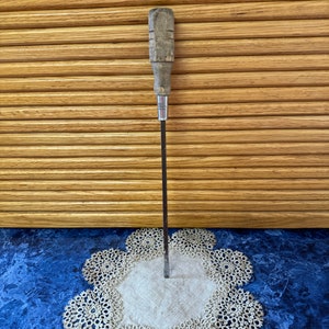 May include: A vintage screwdriver with a wooden handle and a metal shaft. The screwdriver is standing upright on a white doily on a blue surface.