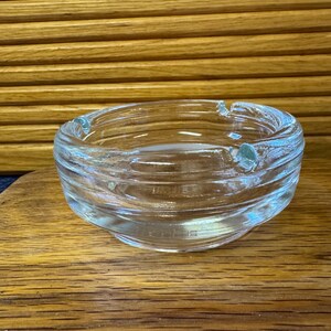May include: Clear glass ashtray with a circular design. The ashtray has a wide, shallow bowl with a series of concentric rings and three small notches for resting cigarettes. The glass has a slight iridescent quality.