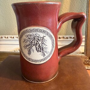 May include: A tall, cylindrical ceramic mug with a deep reddish-brown glaze and a large, curved handle. A circular white medallion on the front features a black line drawing of a pinecone and needles, surrounded by a decorative braided border.