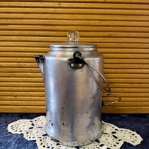 May include: A vintage aluminum coffee pot with a glass knob on the lid and a black handle. The pot sits on a white lace doily against a blue surface. The background is a wooden slatted wall.