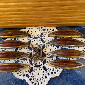 May include: Eight vintage wooden drawer pulls with brass-toned metal trim. The pulls have a dark wood center and are arranged on a white lace doily. A small pile of nails is also present.