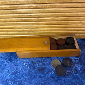 May include: A wooden box with a sliding lid, containing a set of game pieces. The pieces are circular, in shades of brown and black. Three additional pieces are outside the box on a blue surface. The background features a wooden panel.