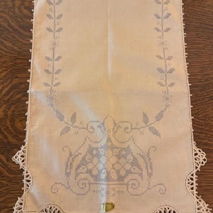 May include: A white linen table runner with a delicate lace border and embroidered floral design. The runner features a central motif of two birds perched on a branch with flowers.