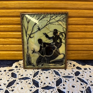 May include: A framed silhouette art piece depicting two figures in a winter scene. The black silhouettes are set against a pale green background, framed in a wooden border. The artwork features a tree and a couple in a horse-drawn sleigh.