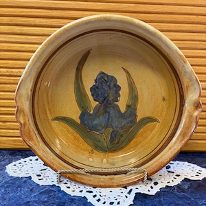 May include: A round ceramic bowl with a tan, speckled glaze and a hand-painted blue iris flower with green leaves in the center. The bowl features brown circular rim accents and scalloped edges on the sides, with the initials JD near the base.