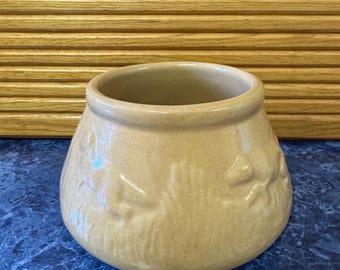 May include: A tan-colored ceramic pot with a rounded base and a wide opening. The pot has embossed floral designs around its exterior. The background features a wooden shutter and a blue countertop.