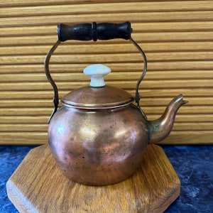 May include: A small, antique copper teapot with a dark wooden handle and a white ceramic knob on the lid. The teapot sits on a wooden hexagonal trivet. The spout and handle are made of brass.
