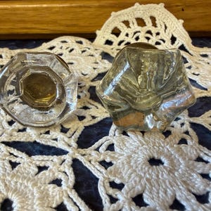 May include: Two clear glass cabinet knobs with a faceted, multi-sided geometric shape. Each knob features a central metallic base component visible through the transparent glass, showcasing a reflective surface and a distinct hexagonal profile.