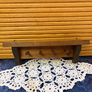 May include: A small, dark-stained wooden shelf with three pegs below. The shelf has a simple design and is mounted on a wooden background. A white lace doily is in the foreground.