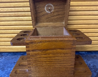 May include: A wooden tobacco pipe stand and storage box. The box is made of light brown wood and has a hinged lid. It features multiple circular holes for holding pipes and a lower tray with additional holes. The box is open, revealing the interior.