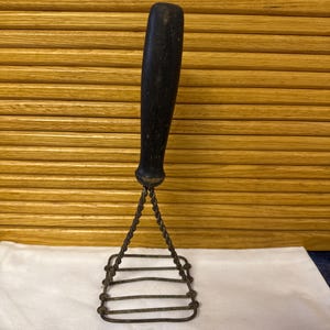 May include: An antique potato masher with a dark wooden handle and a metal mashing head. The handle is cylindrical and appears worn. The mashing head is rectangular with horizontal bars. The masher is resting on a white cloth.