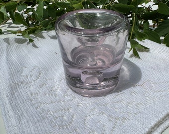 May include: A clear glass candle holder with a purple tint. The holder has a round base and a wide opening at the top.